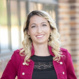 young woman with long blonde hair, wearing purple jacket and a black shirt, smiling at camera. standing in front of a brick building