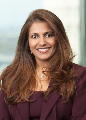Woman with long brown hair wearing a dark red color jacket and sitting in front of a window, smiling at camera